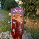 Hlava akustické kytary v tmavě červené barvě s chromovanými ladičkami. V horní části je bílý nápis Squier by Fender®. Povrch lakovaného dřeva vykazuj…
