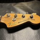 Fotografie zachicuje hlavu basové kytary z přírodního dřeva. Na povrchu je černý nápis Fender a MUSICMASTER BASS, pod kterým je menší text MADE IN US…