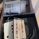 V černé krabičce s průhledným víkem se nachází bílý snímač k elektrické kytáře. Snímač má dvě řady magnetických pólů a je zčásti omotaný černou izola…
