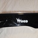 Detail černé lakované části hudebního nástroje s lesklým povrchem. Uprostřed je bílý nápis WOOD v doprovodu grafického prvku připomínajícího blesk. P…