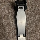Fotografie zobrazuje bicí pedál s kovovou stlačnou deskou stříbrné barvy a černým základním mechanismem. Na spodní části desky je vyražen nápis Rolan…