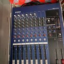 Modrý mixovací pult s nápisem MIXING CONSOLE MG12/4 a logem YAMAHA. Zařízení obsahuje několik vstupních kanálů s ovládacími knoflíky a posuvníky. Pov…