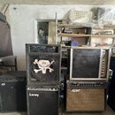 Fotografie zobrazuje čtyři basové komba a zesilovače v interiéru. Vlevo stojí černé kombo s logem AMPEG, vedle něj černý stack s nápisem BASS AMPLIFI…
