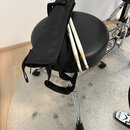 Na černé polstrované židli leží černý textilní obal na paličky a dvě bílé paličky. Na jedné z paliček je čitelný nápis „VIC FIRTH“. Konce paliček vyk…