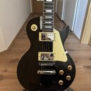 Černá elektrická kytara s tvarem singlecut a krémovým pickguardem. Nástroj má dva humbuckery s kovovými kryty, čtyři zlaté ovládací knofíky a jeden p…