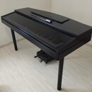 Černý digitální piano v lesklém provedení s uzavřeným víkem. Nástroj stojí na dvou černých nohách a v dolní části jsou vidět pedály. Na horní desce l…