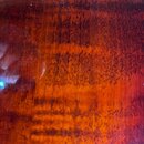 Detailní záběr na povrch hudebního nástroje z reddish-brown dřeva s výraznou masivní strukturou slupů a žil. Povrch je lakovaný s vysokým leskem, na…