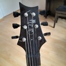 Fotografie zachycuje hlavu elektrické kytary v černé barvě s kovovými ladičkami. Na povrchu je bílý nápis "Paul Reed Smith SE" doplněný o podpis auto…