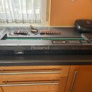 Černý digitální klavír značky Roland, který je rozložený na pracovní desce a nemá kryt klávesnice. Na zadní straně je vidět bílý nápis Roland a štítk…