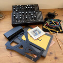 Na dřevěném stole je černý analogový syntetizátor s nápisy MOTHER-32 a moog. Vedle něj leží černý audio interface s nápisem CORAL, sada barevných pat…