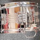 Chromovaný snare drum s kovovým korpusem a stříbrným povrchem. Na nástroji jsou vidět ladicí šrouby, napínací mechanismy a úchyt s madlem. Povrch je…
