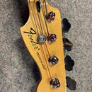 Hlava basové kytary z přírodního dřeva s четыma chromovanými ladičkami. Na povrchu je černý nápis Fender® PRECISION BASS®. Povrch dřeva vykazuje drob…