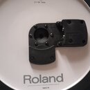 Bílý elektronický činel s černým gumovým okrajem a černým plastovým úchytem v centru. Na povrchu jsou nápisy V-Drums, CY-13R Ride a Roland. Dole je u…