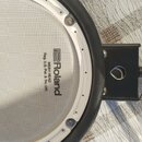 Elektronický buben s bílou pletenou membránou a černým rámem. Na povrchu je černý nápis Roland, MESH HEAD a Reg. U.S. Pat. & Tm. Off., orazvně s loge…