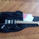 Černá elektrická kytara s bílým hmatníkem a černým pickguardem, položená na černém pouzdře. Na hlavě kytary je viditelné logo Fender a nápis STRATOCA…