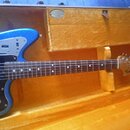 Modrá elektrická kytara s kovovými prvky a černým pickguardem v žlutém kufríku. Na hlavě kytary je nápis Fender Jaguar. Hриф má hnědý prstový hmatník…