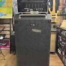 Černý basový reproduktor v bedně na kolečkách s kovovou rukojetí. V horní části je umístěn otevřený hliníkový flightcase, ve kterém se nachází zesilo…