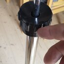 Detailní záběr na část chromovaného stojanu s černým plastovým šroubovacím knoflíkem. Nad knoflíkem je vidět kovová tyč a šedý plstěný nebo textilní…