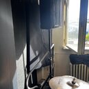 Černý aktivní reproduktor umístěný na černém stativu. V dolní části je vidět část činely s nápisem Zildjian. V pozadí je okno a bílý radiátor.