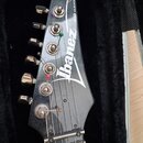 Detail hlavy elektrické kytary v černé barvě, která leží v černém pouzdře. Na hlavě je bílé logo Ibanez a svislý nápis SERIES. Jsou vidět šest kovový…