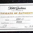 Fotografie znázorňuje bílý papírový certifikát s nápisem CERTIFICATE OF AUTHENTICITY a logem Xotic Guitars CALIFORNIA. Dokument obsahuje text „This d…