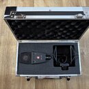 Otevřený hliníkový kufr s černou pěnovou výplní, ve kterém je umístěn černý dynamický mikrofon a držák proti rázům (shock mount). Na těle mikrofonu j…