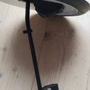 Černý kovový stojan s úchytem, který drží stříbrný kulatý disk. Na horní části disku je viditelný nečitelný nápis. Celý předmět leží na světlém dřevě…