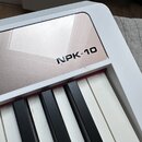 Detail bílého klávesového nástroje s černými a bílými klávesami. Na horní části je stříbrný panel s černým nápisem NPK-10. Povrch nástroje vykazuje d…