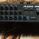 Zadní strana černého modulu elektronických bicích s nápisem ALESIS STRIKE. Jsou zde vidět početné vstupy a výstupy, včetně MIDI OUT, MIDI IN, AUX IN…