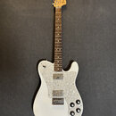 Bílá elektrická kytara s hnědým hmatníkem a perleťovým pickguardem. Na hlavě je viditelné logo Fender a nápis Telecaster Deluxe. Nástroj vykazuje zná…
