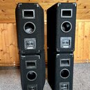 Čtyři černé reproduktorové boxy v provedení 2x2. Každý box má v horní části hranolitý otvor a uprostřed kulatý odvětrací otvor s mřížkou. Na spodní č…