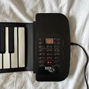 Černý elektronický klavír s bílými a černými klávesami. Na ovládacím panelu je digitální displej s červenými číslicemi 3.8.8 a nápis ROCK ROLL IT! s…