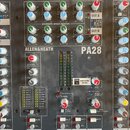 Detailní pohled na míchací pult značky ALLEN&HEATH, model PA28. Povrch je šedý s množstvím bílých a barevných ovratových knoflíků, posuvníků a červen…