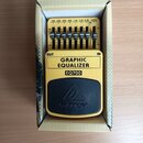 Žlutý kytarový efekt v kartonové krabici. Na černém horním krytu je logo behringer a uprostřed trojúhelníkový symbol. Pod ním je nápis EQ700 a štítek…