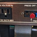 Detail zadní strany zařízení s černým povrchem a bílými popisky. Vlevo je napájecí konektor s nápisy 120 VOLTS 60 Hz a 300 WATTS MAX. Vpravo se nachá…