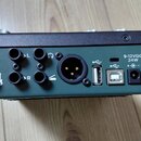 Zadávací panel černého zařízení s tmavě zeleným pozadím. Jsou zde vidět vstupy a výstupy s nápisy L, R, symboly sluchátek a kytary, USB porty a konek…