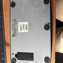 Fotografie zachycuje spodní stranu černého elektronického zařízení s kovovým dnem. Na spodní části jsou čtyři kulaté gumové nožičky a dvě nálepy s te…