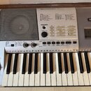 Stříbrošedý elektronický klavír značky YAMAHA. Na horním panelu jsou tabulky s názvy SONG a STYLE, ovládací knoflík MASTER VOLUME a několik tlačítek,…