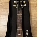 Kytara s tmavě hnědým hmatníkem a černou hlavou v černém obalu. Na hlavě je bílé kulaté logo s hvězdicí a na obalu nápis YAMAHA doprovázený stejným l…