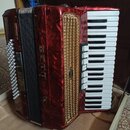 Červená akordeon s klaviatura a zlatou mřížkou. Na boku je svisle napsaný bílý nápis DELI a pod ním písmeno V. Na zlaté mřížce je malý oválný štítek…