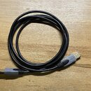 Černý USB kabel s šedými konektory, svinutý do kruhu na dřevěném povrchu. Na jednom z konektorů je vyryto logo USB. Ve spodní části fotky je vidět čá…