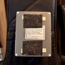 Štihlá šedá kovová krabička s čtyřmi viditelnými šrouby v rozích. Na povrchu jsou dva černé suché zapínací pásy a bílá nálepka s textem: „subject to…
