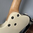 Detail zadní strany kytary v místě spoje krku a těla. Tělo je v krémové barvě s čtyřmi viditelnými šrouby u základny hmatu, krk je z přírodního dřeva…