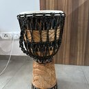 Dřevěný buben s bílou membránou a černým pleteným provázkem s barevnými detaily. Tělo nástroje je zdobeno vyřezávanými ornamenty a tmavým povrchovým…