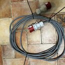 Šedý silový kabel smouseDowným červeným konektorem a zásuvkou. Na bílém plastovém krytu konektoru jsou nečitelné nápisy. Kabel je svázaný černými pla…