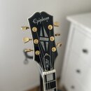 Fotografie zachycuje hlavu elektrické kytary v černé barvě s bílým lemováním. V horní části je bílé logo Epiphone a níže nápis Les Paul CUSTOM. Nástr…