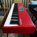 Červený digitální klavír s bílými a černými klávesami. Na horním panelu je bílý nápis nord piano a ovládací sekce s knoflíky a tlačítky. Povrch červe…