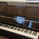 Hnědé pianino s vysokým leskem a viditnou strukturou dřeva. Na krytu nad klávesnicí je zlatý nápis AUGUST FORSTER. Klávesnice je v bílo-černém proved…