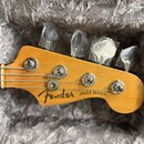 Fotografie zachicuje hlavu basové kytary v přírodním dřevě, která leží v šedém plyšovém pouzdře. Na hlavě je černý nápis Fender a JAZZ BASS. Ladicí k…