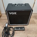 Černý kytarový kombozesilovač s bílým rámečkem kolem mřížky a logem VOX. Na horním ovládacím panelu jsou umístěny ovratidla a přepínače. Před zesilov…
